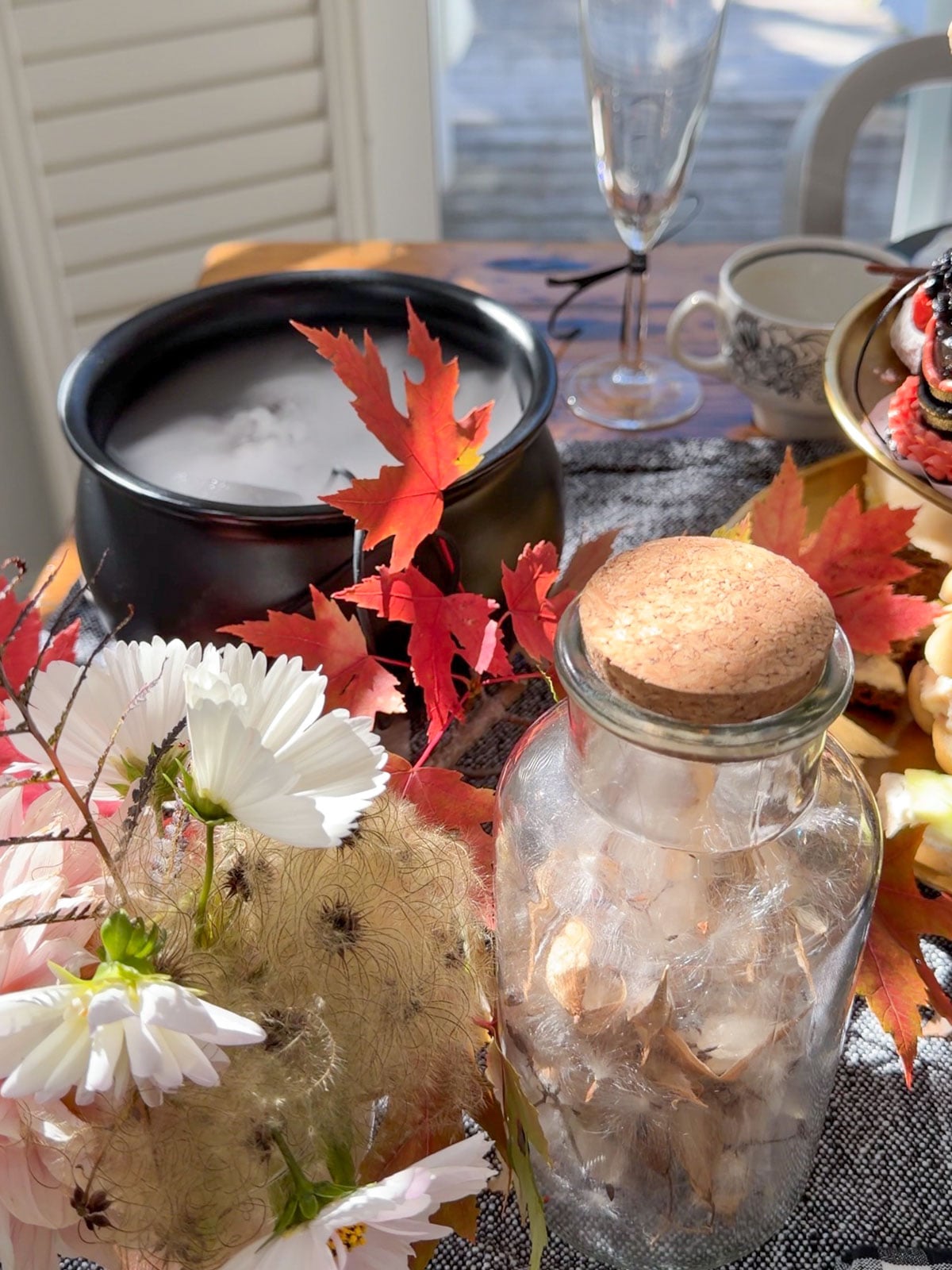 Black cauldron filled with fog beside maple leaves, white cosmos flowers and a glass apothecary jar of dried seed pods.