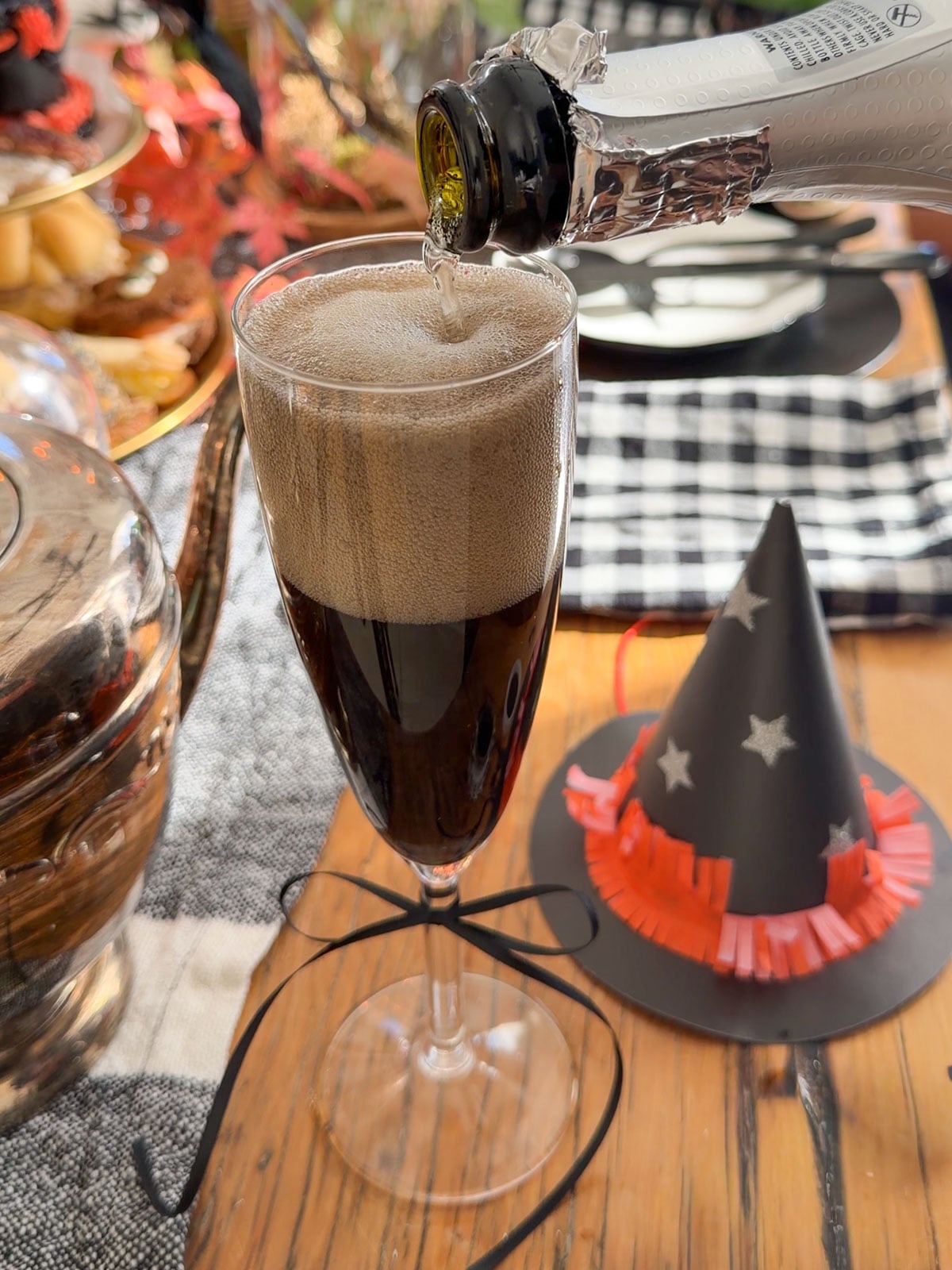 Black Kir Royale witch themed cocktail in a champagne flute tied with a black ribbon, with a paper witch hat beside it.
