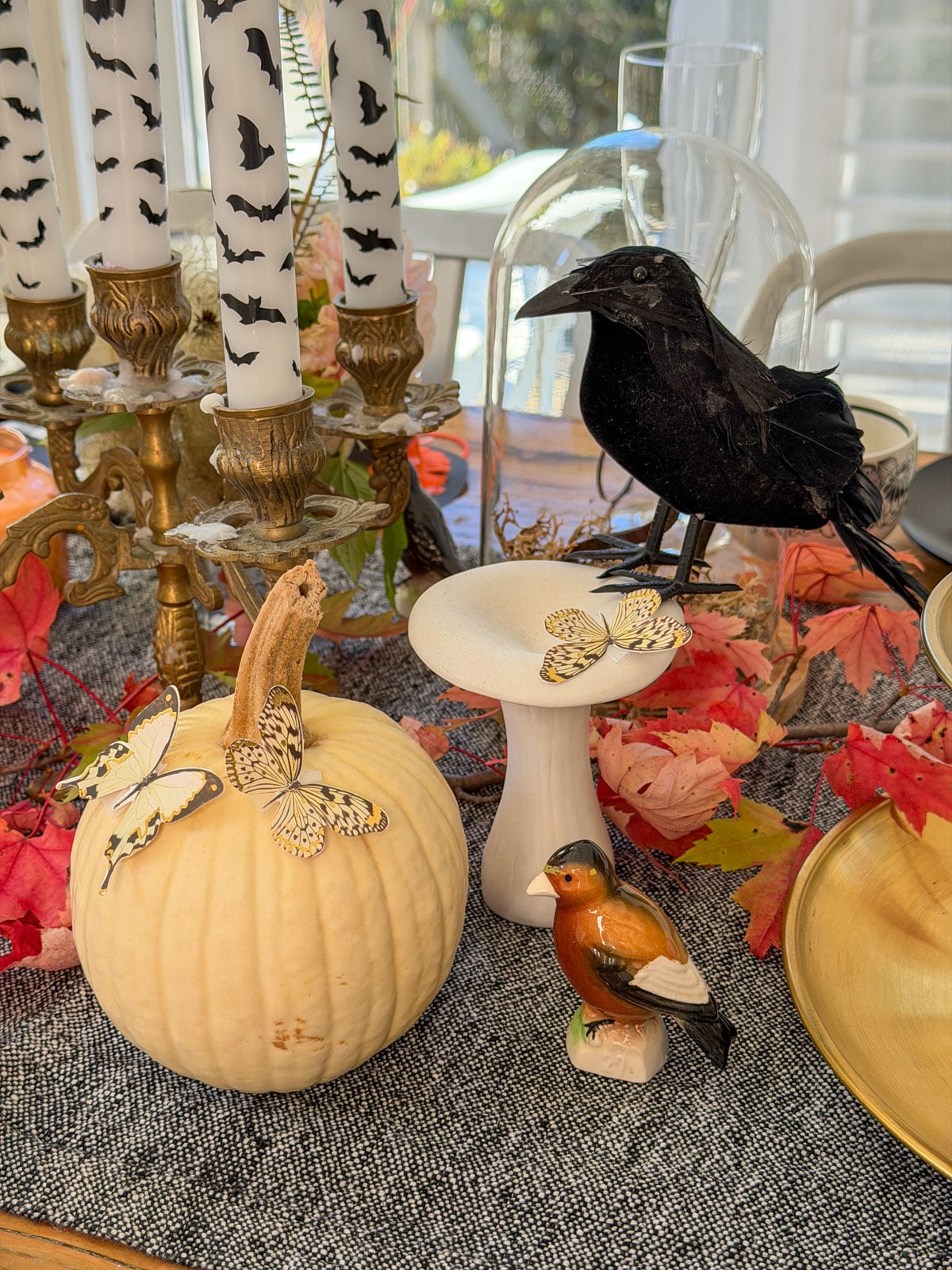 White pumpkin decorated with paper butterflies beside a black crow on a white mushroom stand, a bird figurine and bat candles.